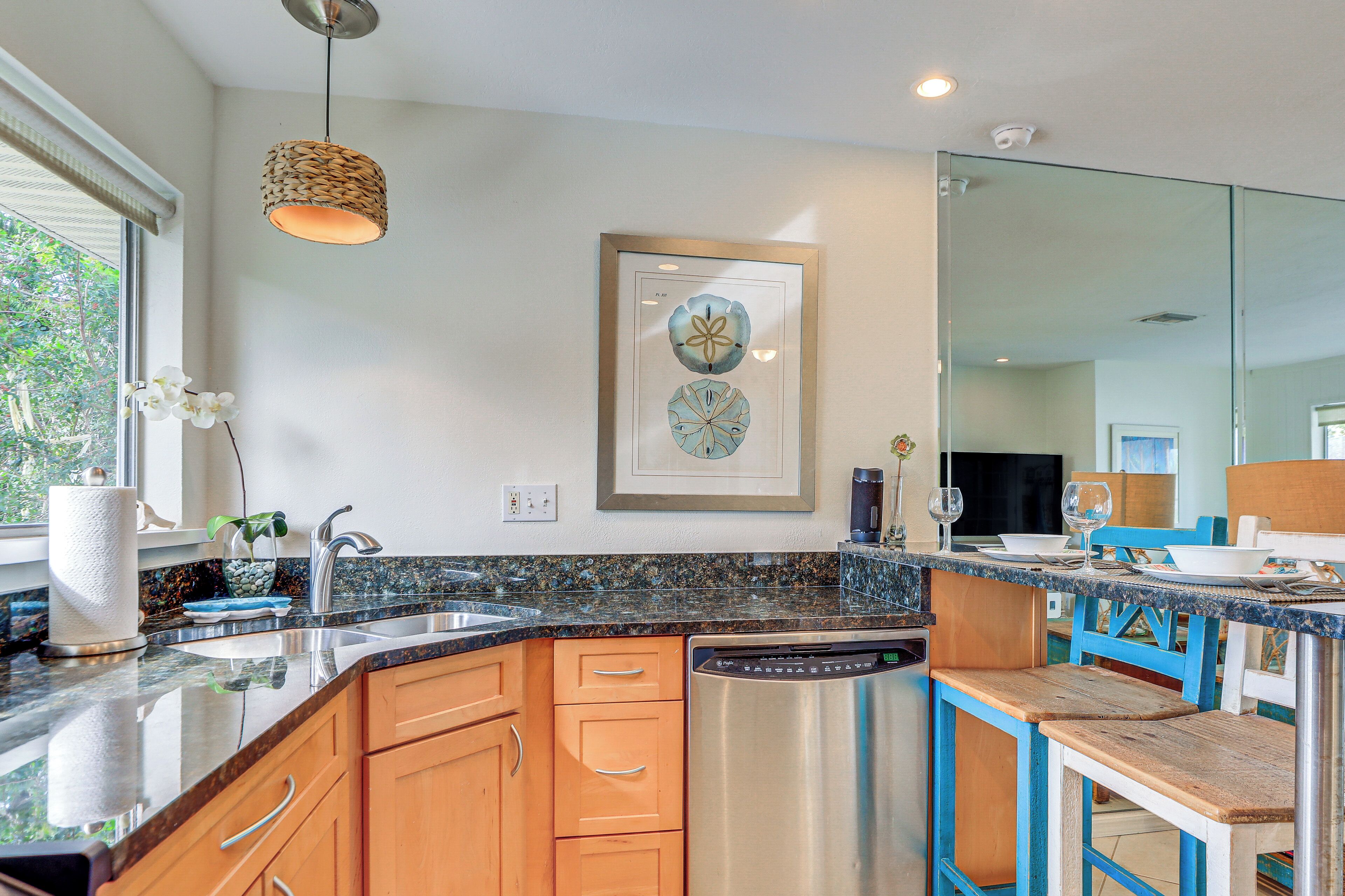 A modern kitchen with granite countertops, wooden cabinets, a dishwasher, and a dining area with blue chairs.