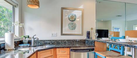 A modern kitchen with granite countertops, wooden cabinets, a dishwasher, and a dining area with blue chairs.
