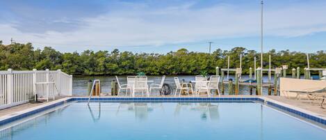 Une piscine entourée de chaises blanches et d’une jetée qui s’avance sur une étendue d’eau, le tout bordé de verdure.
