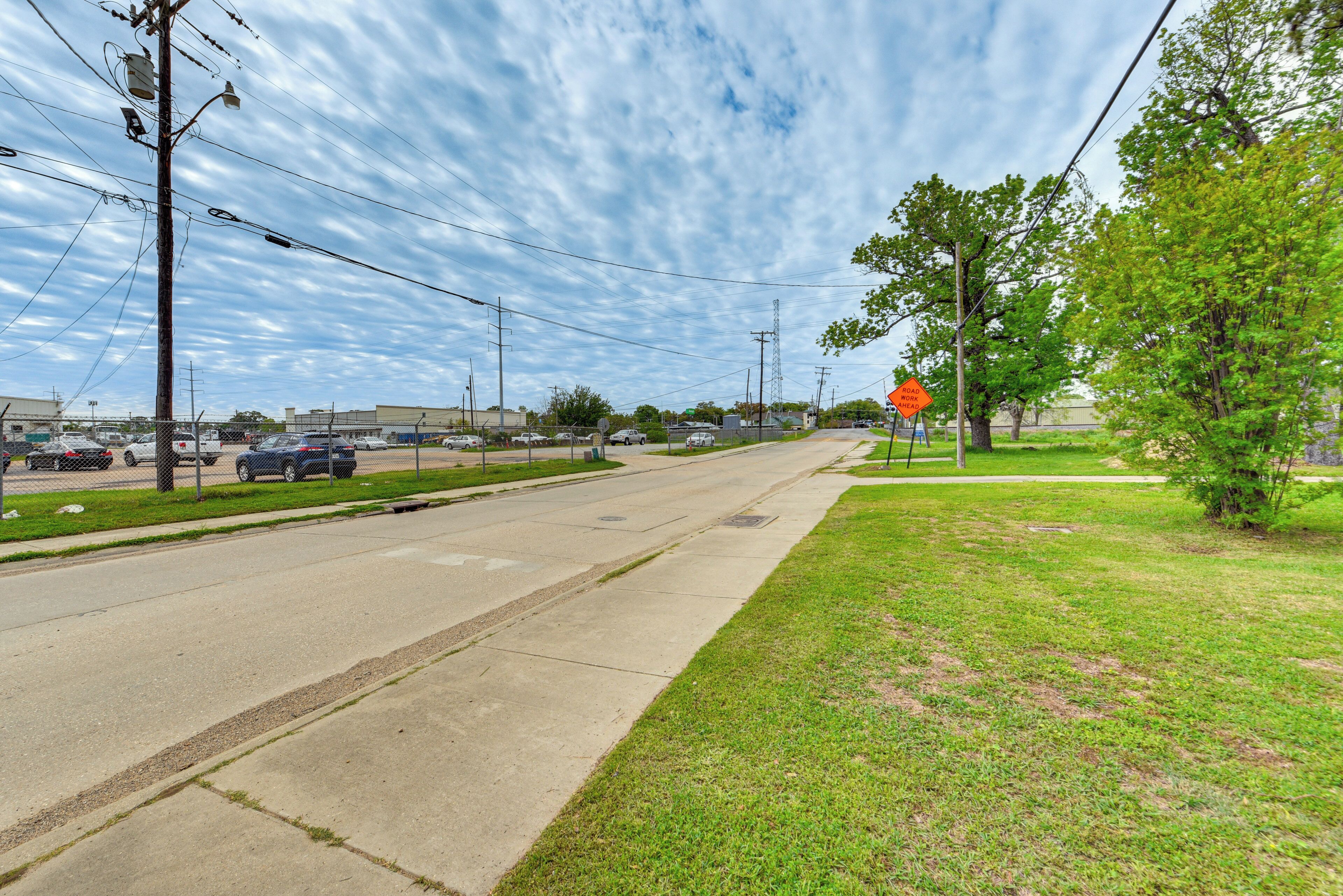 A wide road with a sidewalk on one side and a grassy area on the other. There are power lines and a few buildings in the background.