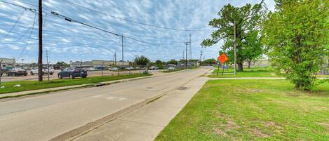 A wide road with a sidewalk on one side and a grassy area on the other. There are power lines and a few buildings in the background.