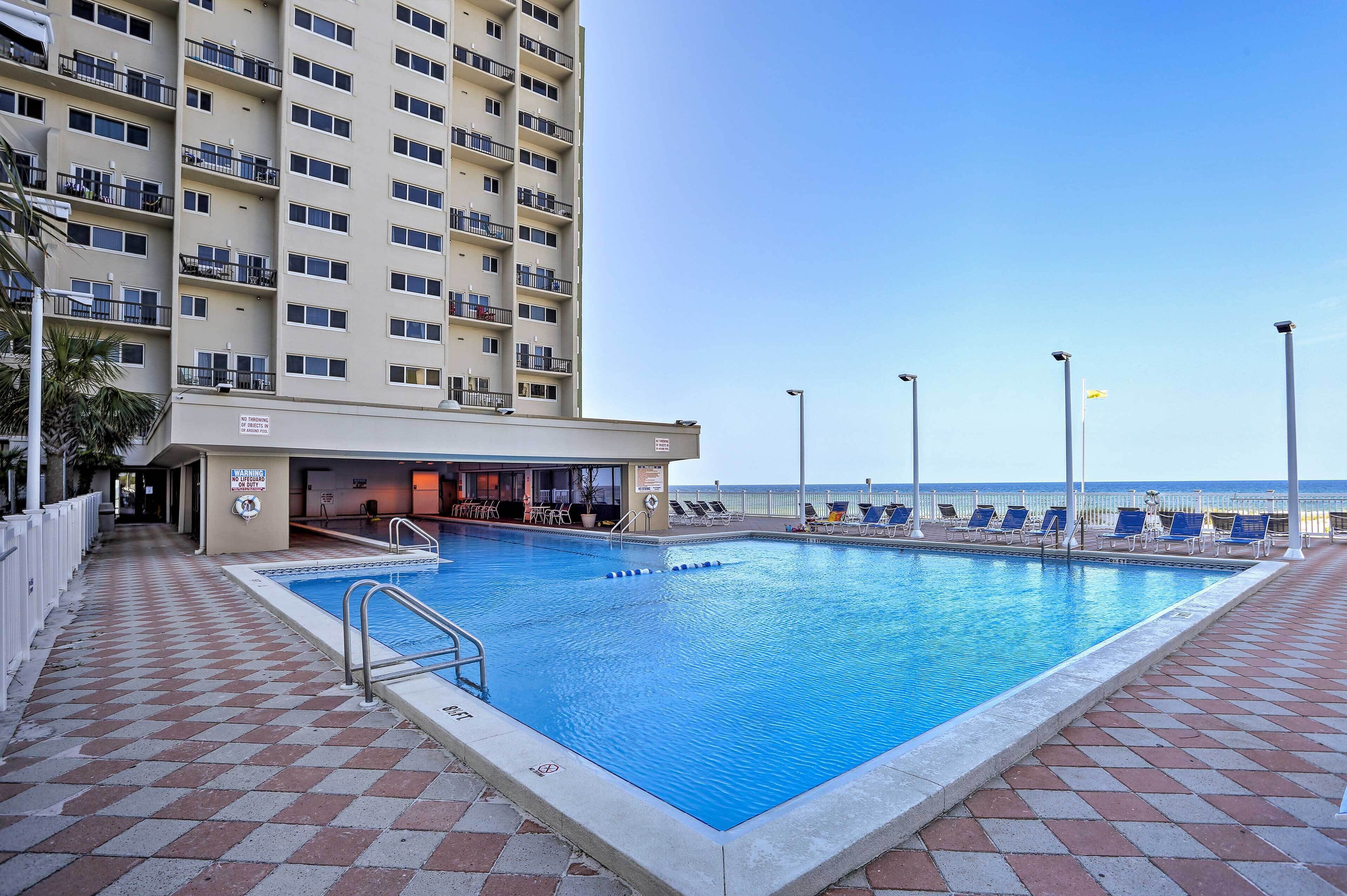 A swimming pool with a tiled deck, a building with balconies, and a view of the ocean.