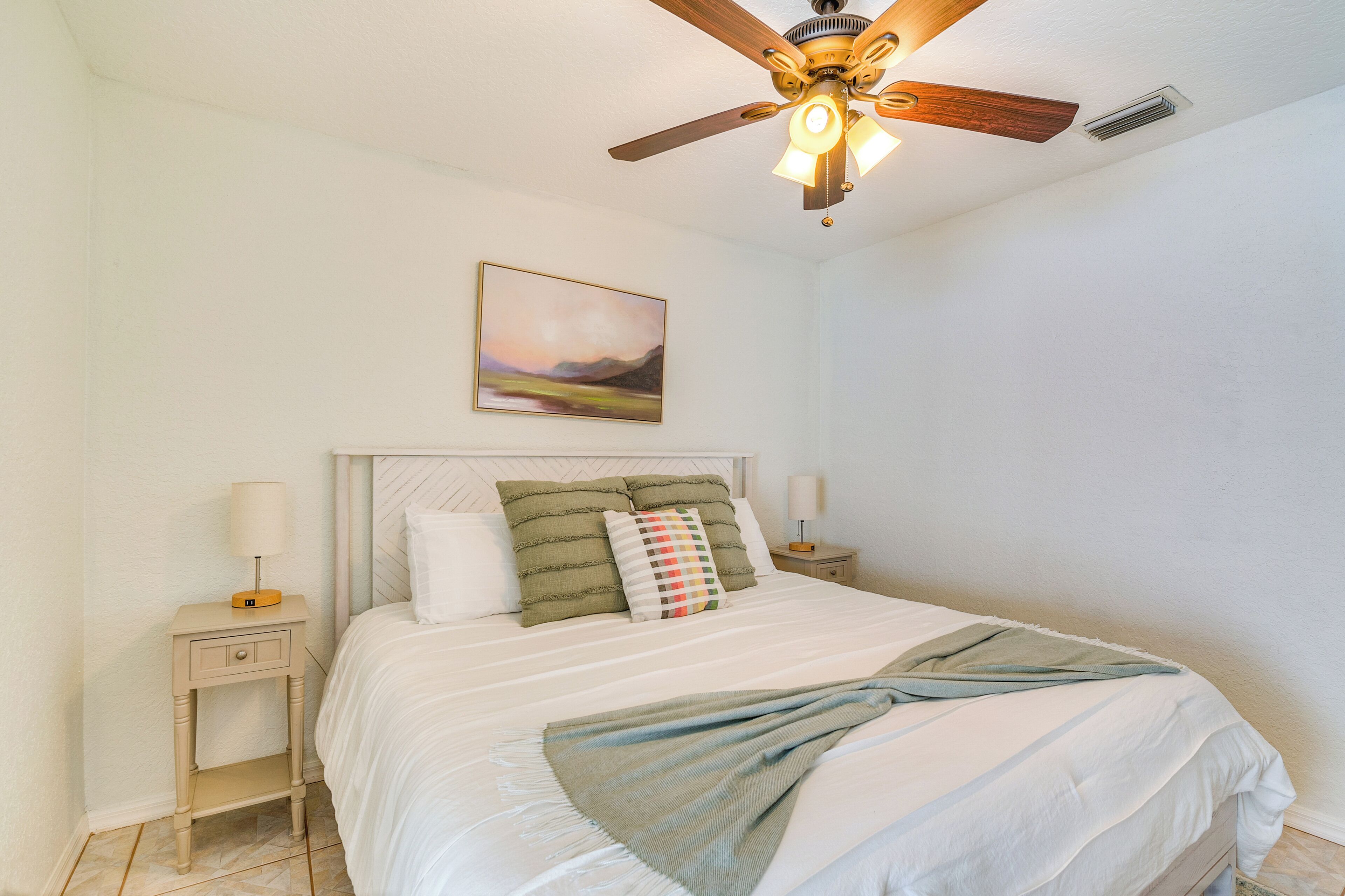 Une chambre à coucher comprenant un lit, une table de chevet, un ventilateur de plafond et un tableau accroché au mur.