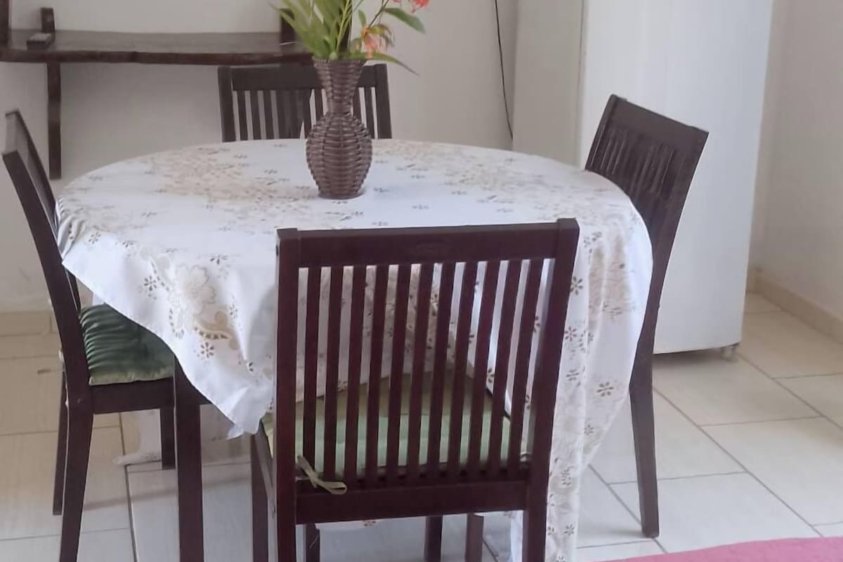 A dining area with a table set for two, a vase with flowers, and a refrigerator in the background.