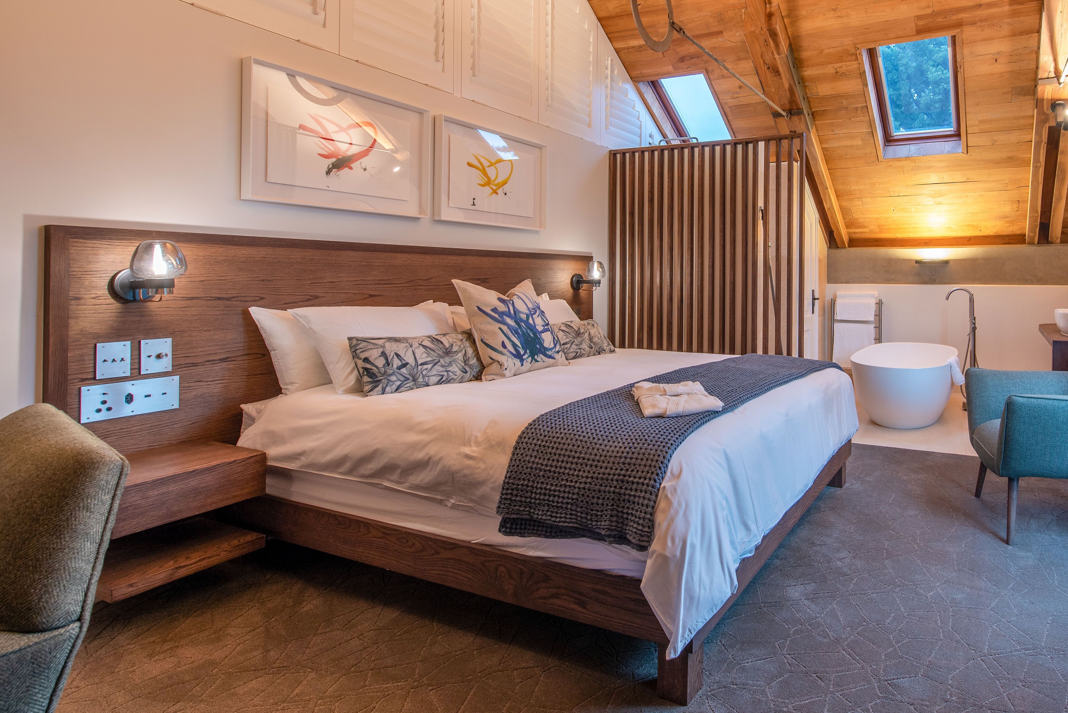 Ein modernes Schlafzimmer mit einem Holzkopfteil, einem großen Bett mit vielen Kissen, einem Nachttisch, einer Wandlampe und einem Oberlicht.
