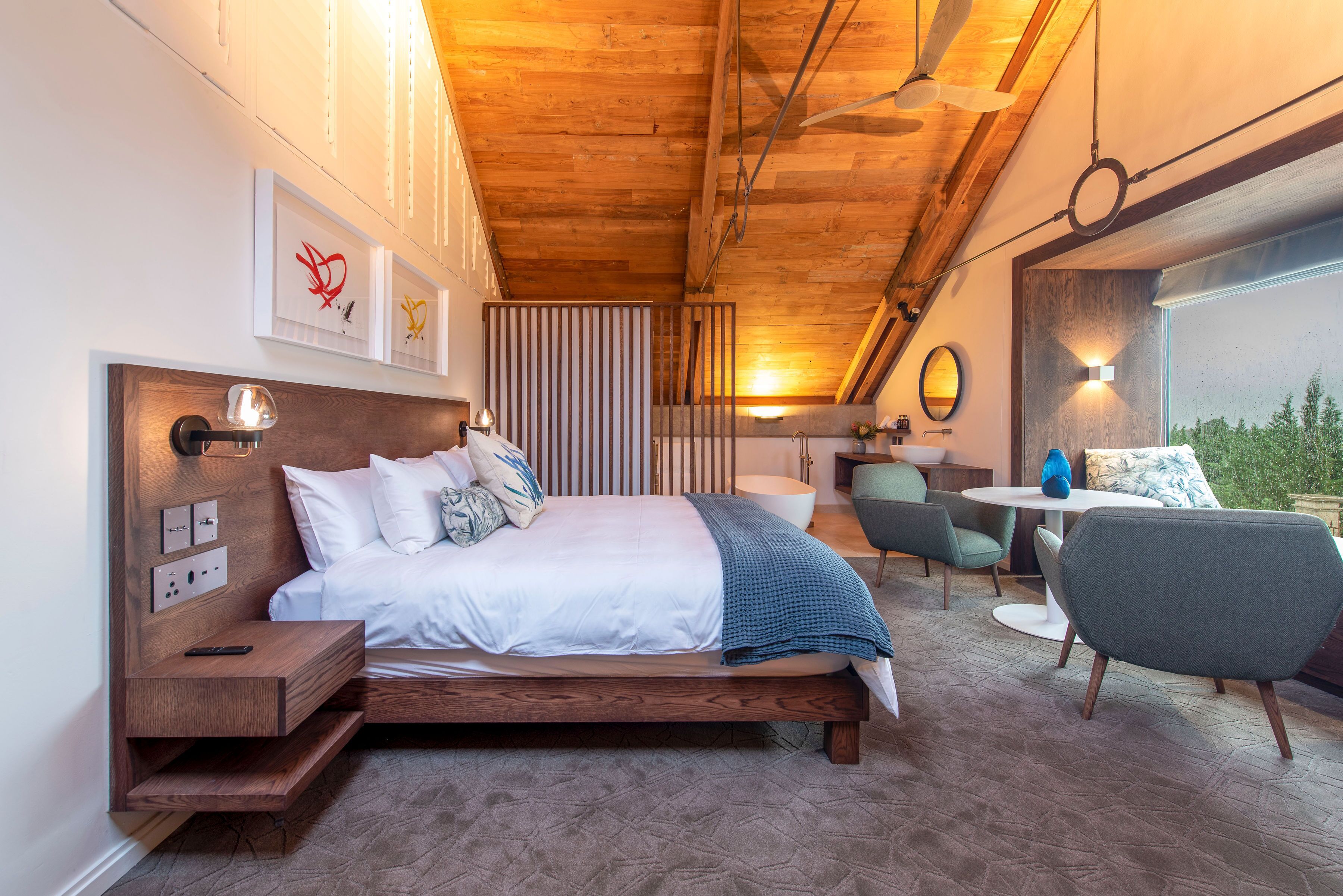 A modern bedroom with a wooden ceiling, a bed with white bedding, a bedside table, a chair, and a small table with a lamp.