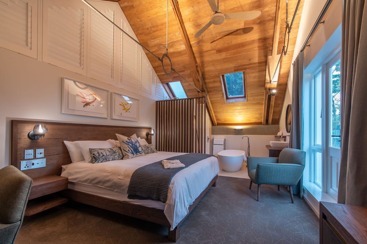 A bedroom with a wooden ceiling, a bed with a headboard, a bedside table, a chair, a window, and a bathroom with a sink and mirror.