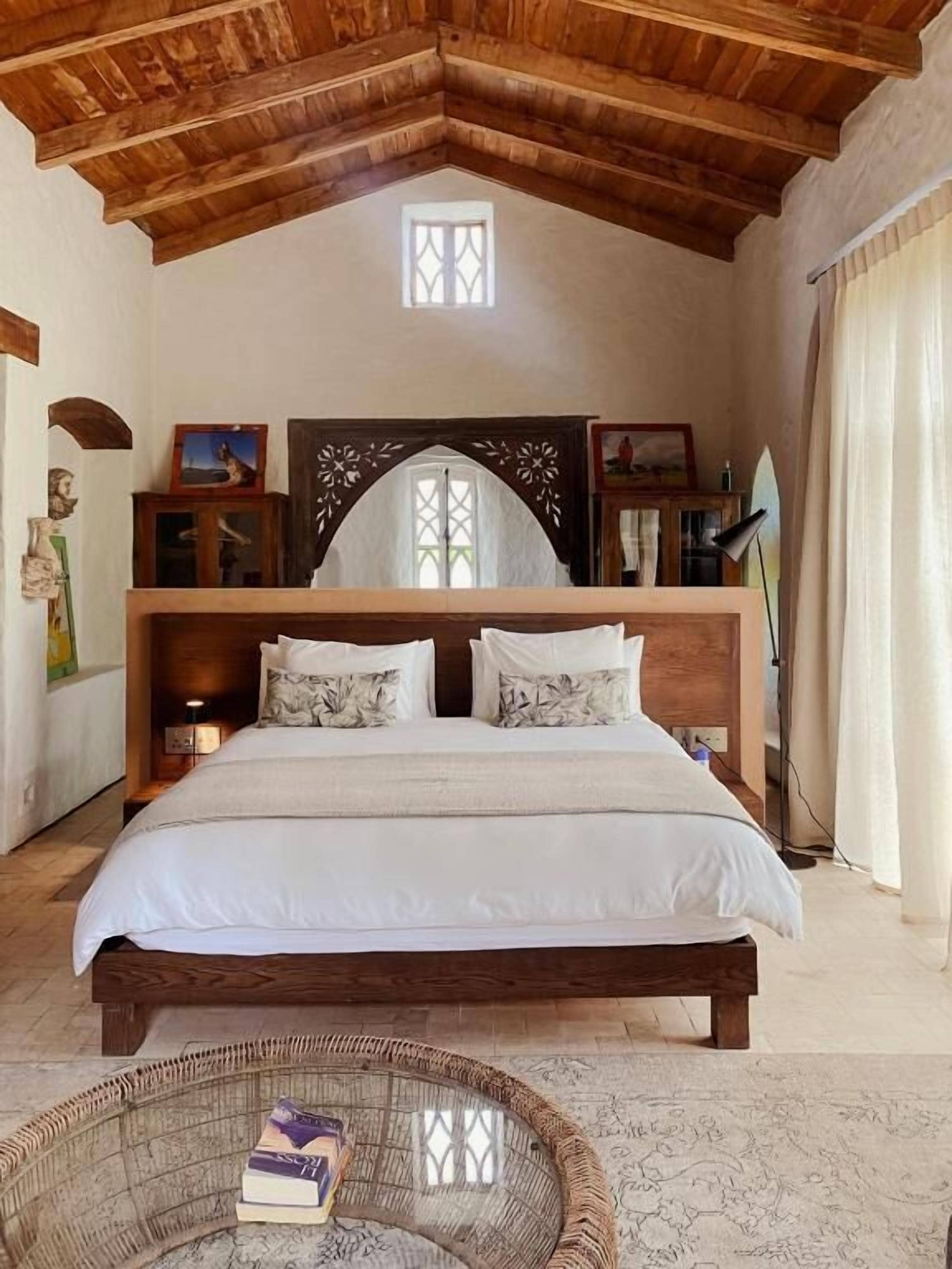 Une chambre avec un plafond en bois, un grand lit avec une literie blanche, une petite fenêtre et une tête de lit décorative en bois.