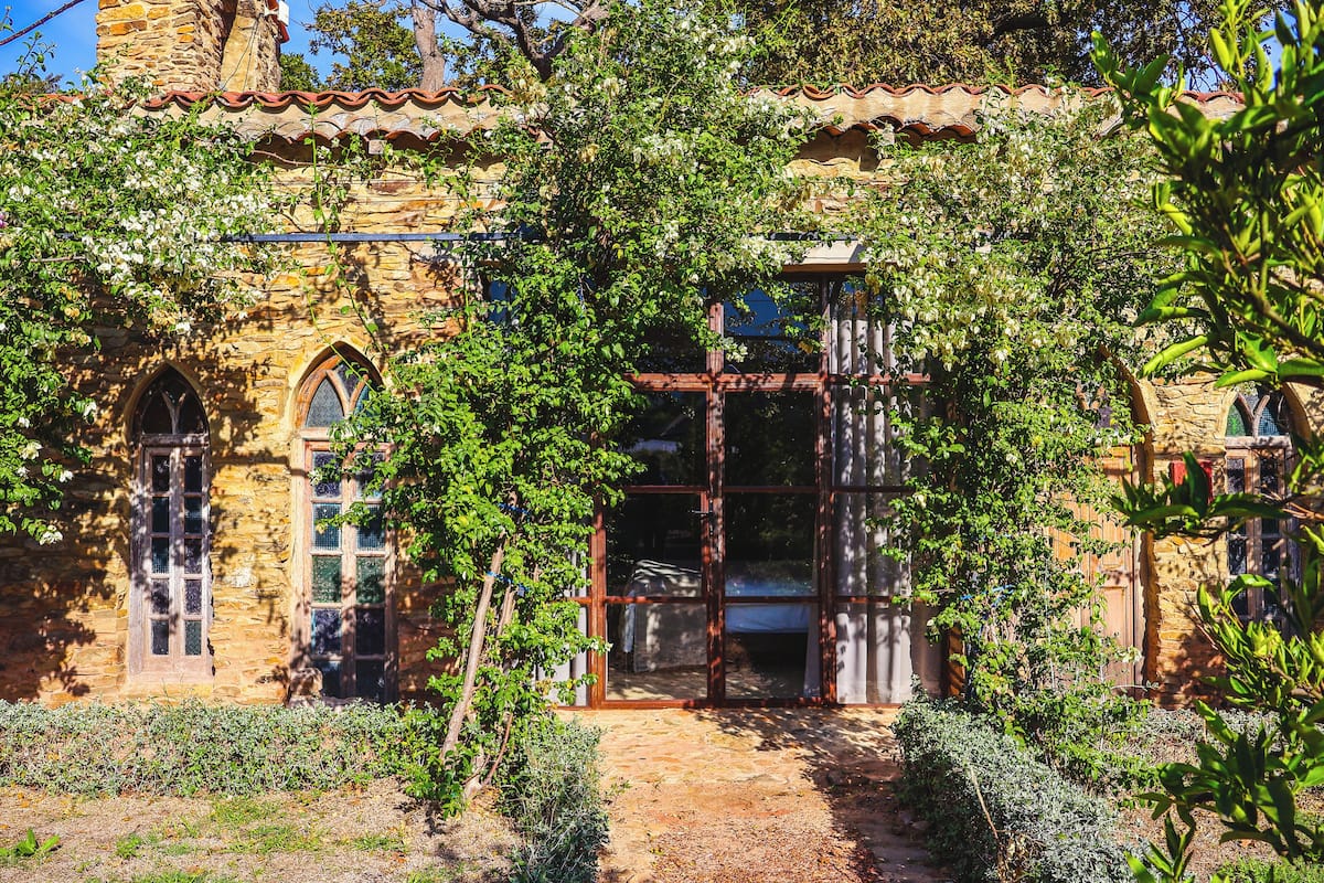 A stone building with arched windows and a wooden door, surrounded by lush greenery.