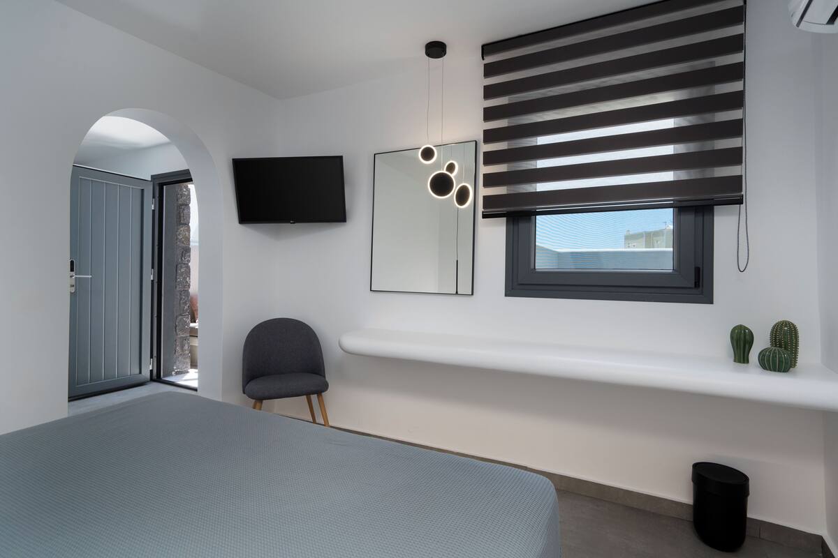 Un dormitorio moderno con una cama, una silla, un televisor, una ventana con persianas y un estante pequeño con adornos.