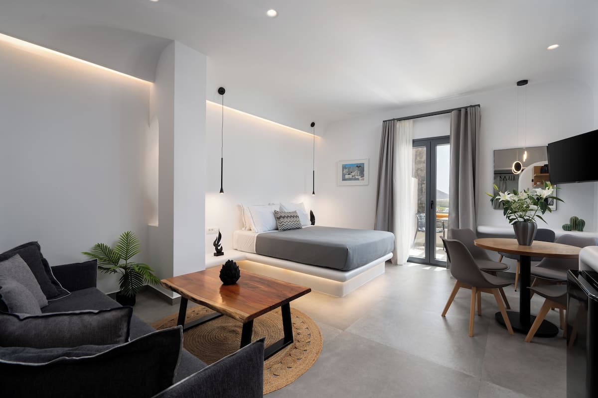 Una sala de estar moderna y bien iluminada con un sofá, una mesa de centro, un comedor con sillas, una cama con colchón gris y un televisor montado en la pared.
