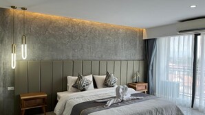 Una habitación de hotel moderna con una cama grande, una mesita de noche de madera, una mesilla auxiliar y vistas al exterior a través de amplias ventanas.