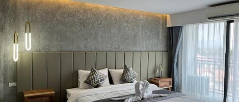 Una habitación de hotel moderna con una cama grande, una mesita de noche de madera, una mesilla auxiliar y vistas al exterior a través de amplias ventanas.