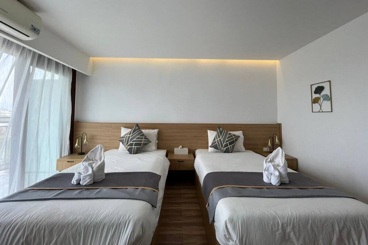 Ein Hotelzimmer mit zwei Betten, einem Holzkopfstück, Nachttischlampen und einem Fenster mit durchsichtigen Vorhängen.
