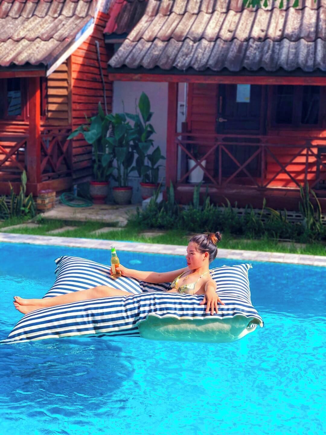 Una persona descansando en una silla de piscina a rayas, con una cabaña de madera y plantas en macetas al fondo.