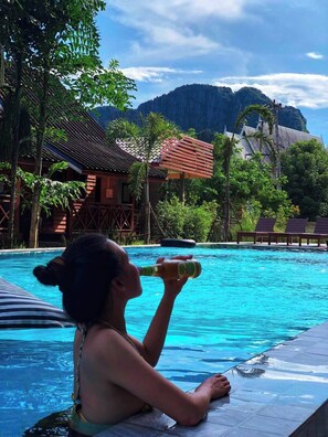 A poolside view with a person relaxing by the pool, surrounded by lush greenery and traditional architecture. - Vang Vieng Remember Hotel (Vang Vieng)