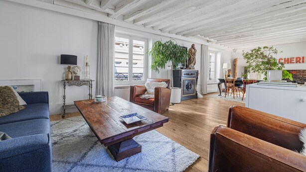 Uma sala de estar ampla, com uma mesa de centro de madeira, sofá azul e uma área de jantar com mesa e cadeiras brancas.