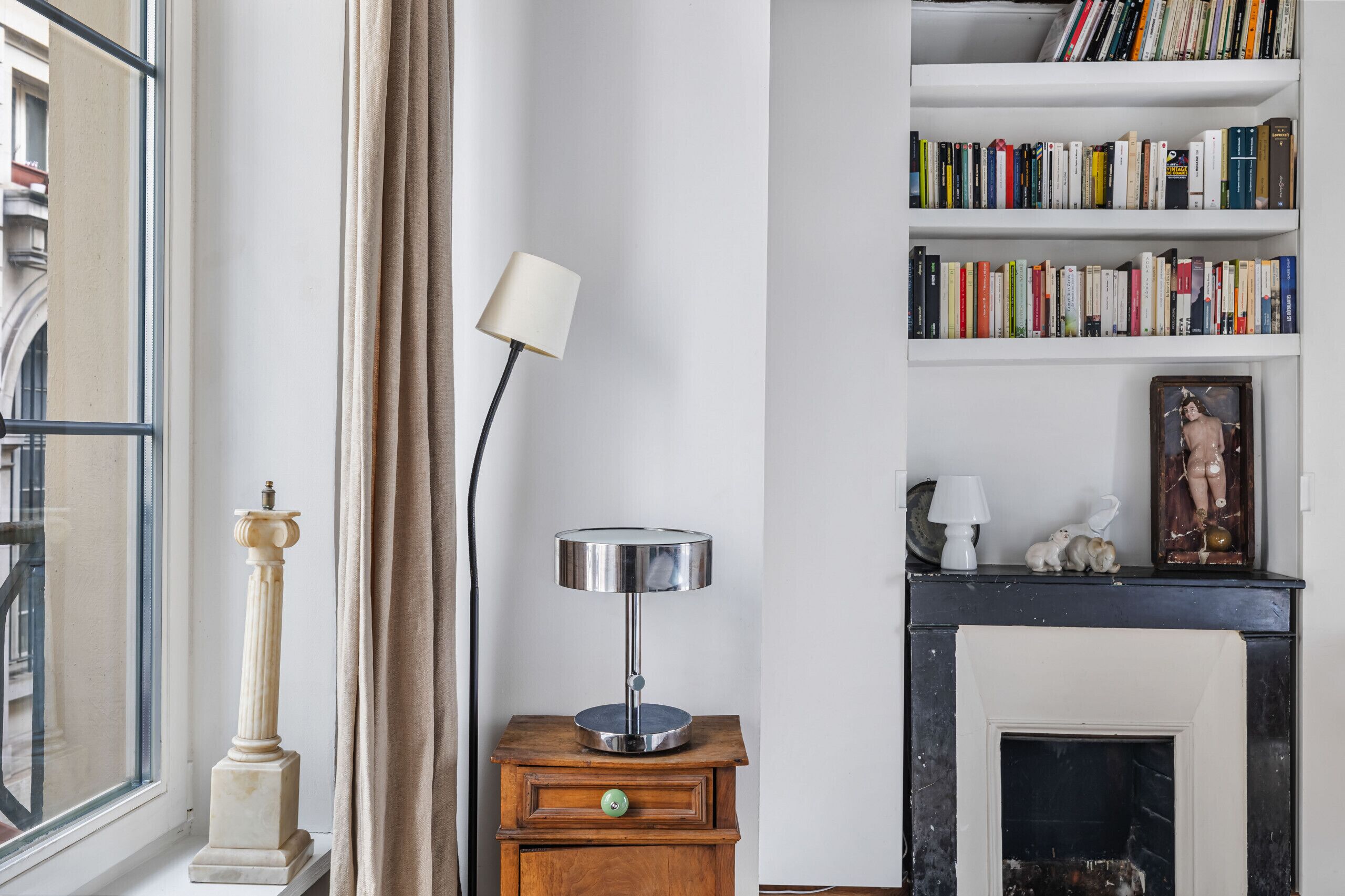 A room with a bookshelf, a lamp, a fireplace, and a window with curtains.