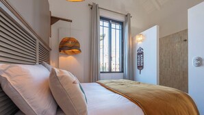 Een slaapkamer met een bed, kussens, een raam en een wandlamp.