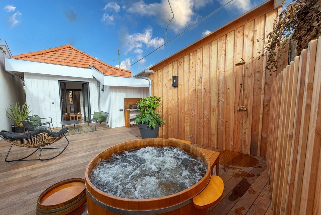 Eine Holzterrasse mit Whirlpool, Sitzgelegenheiten im Freien und einem Gebäude mit rotem Dach.