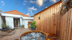 Een houten terras met een jacuzzi, buitenmeubels en een gebouw met een rood dak.