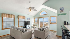 A living room with two sofas, a coffee table, a TV, and a ceiling fan. There are large windows with curtains, a view of the ocean, and a sign on the wall. - Islander: Oceanfront | Hot Tub | Dog-Friendly (Rodanthe)