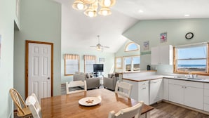 A spacious living room with a wooden dining table, white chairs, a ceiling fan, and a large window offering a view of the ocean. - Islander: Oceanfront | Hot Tub | Dog-Friendly (Rodanthe)