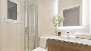 A modern bathroom with a glass shower enclosure, a white toilet, a wooden vanity with a mirror, and a vase with flowers.