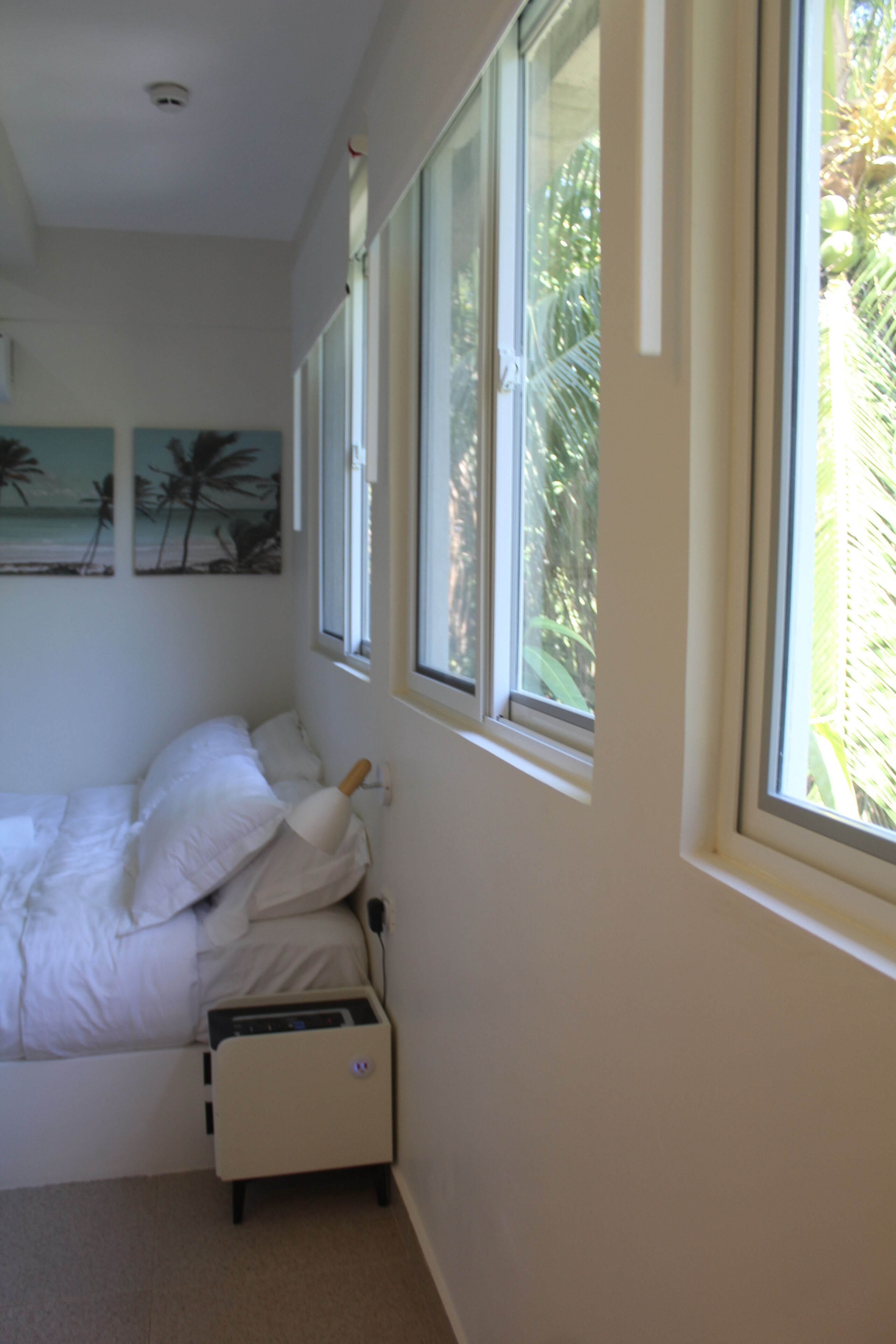 A bedroom with a bed, a bedside table, and a window with a view of palm trees.