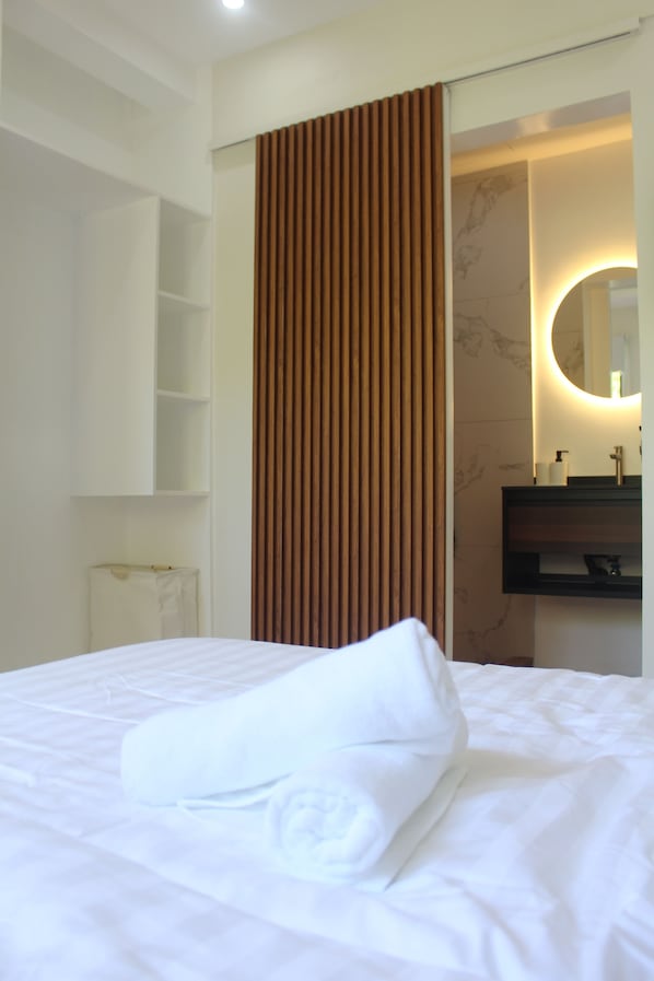 A hotel room with a neatly made bed, a wooden panel wall, a bathroom with a mirror and sink, and a white towel on the bed.