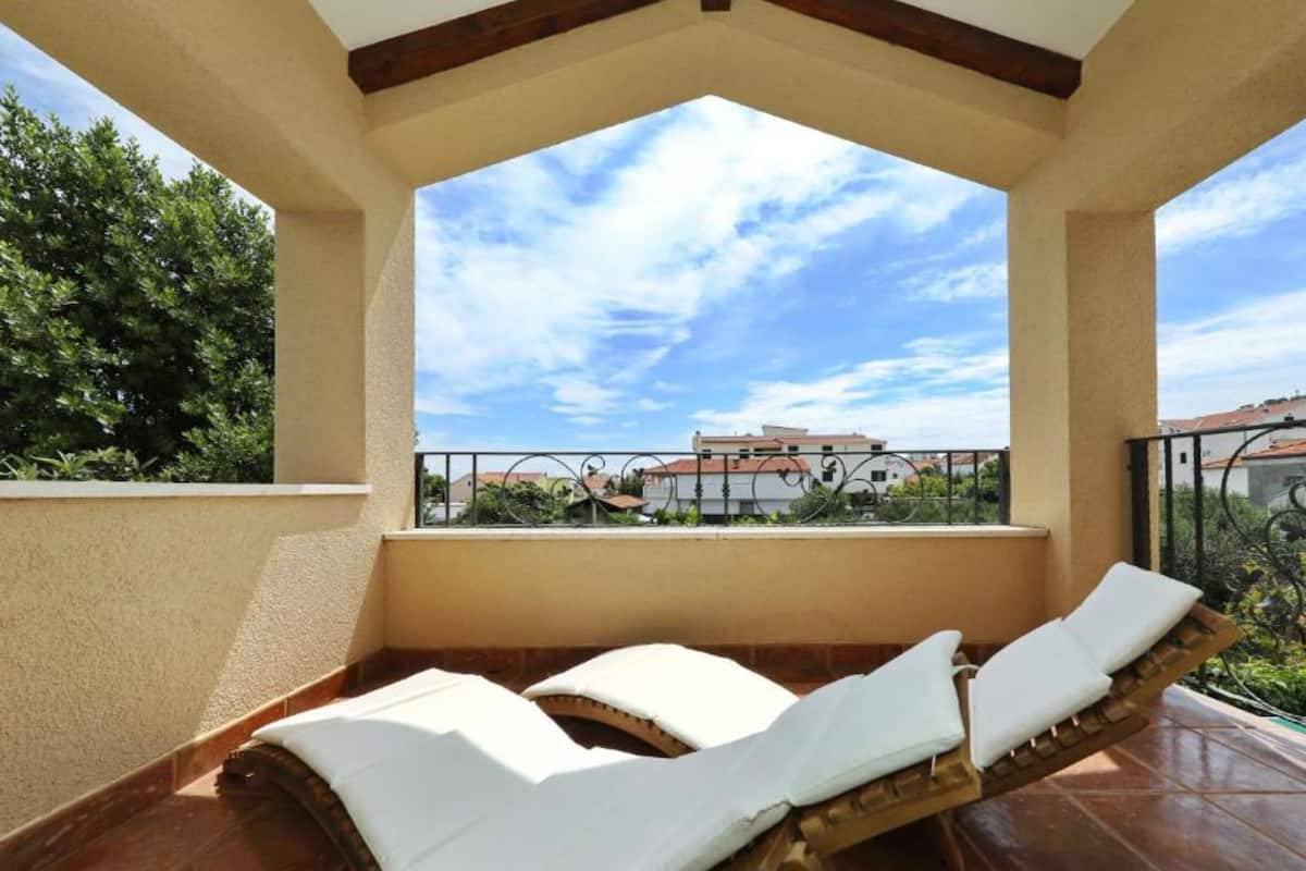 Un balcon avec deux chaises longues, un sol carrelé et une vue sur les bâtiments et les arbres.