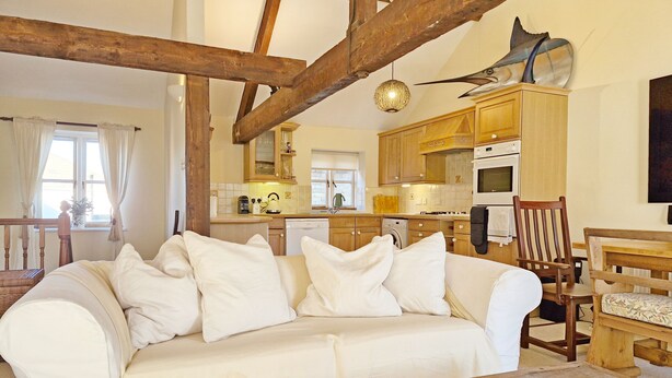 Ein gemütliches Wohnzimmer mit weißem Sofa, Holzbalken, einem Esstisch und einer Küche im Hintergrund.