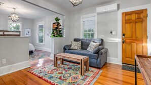 A spacious living room with hardwood flooring, a gray sofa, a wooden coffee table, and a colorful rug. There are two pendant lights, a wooden door, and a window with a view of greenery. - 25% Off Sept, Marble Falls Suite, 1 Block To DT! (Marble Falls)