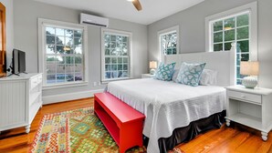 Quarto com cama, um banquinho vermelho, ventilador de teto e janelas amplas.