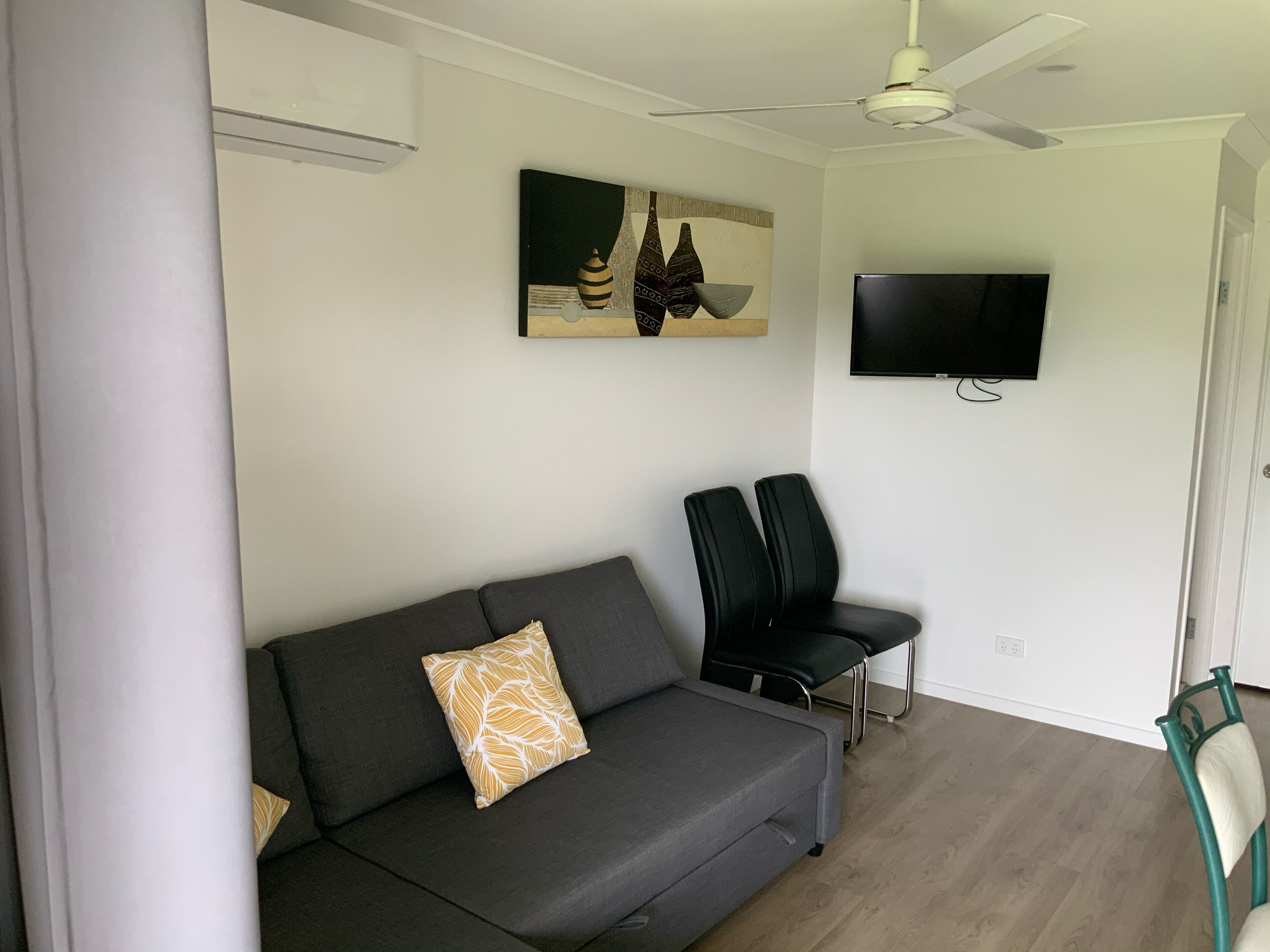 A modern living room with a grey sofa, two black chairs, a ceiling fan, a mounted TV, and a painting on the wall.
