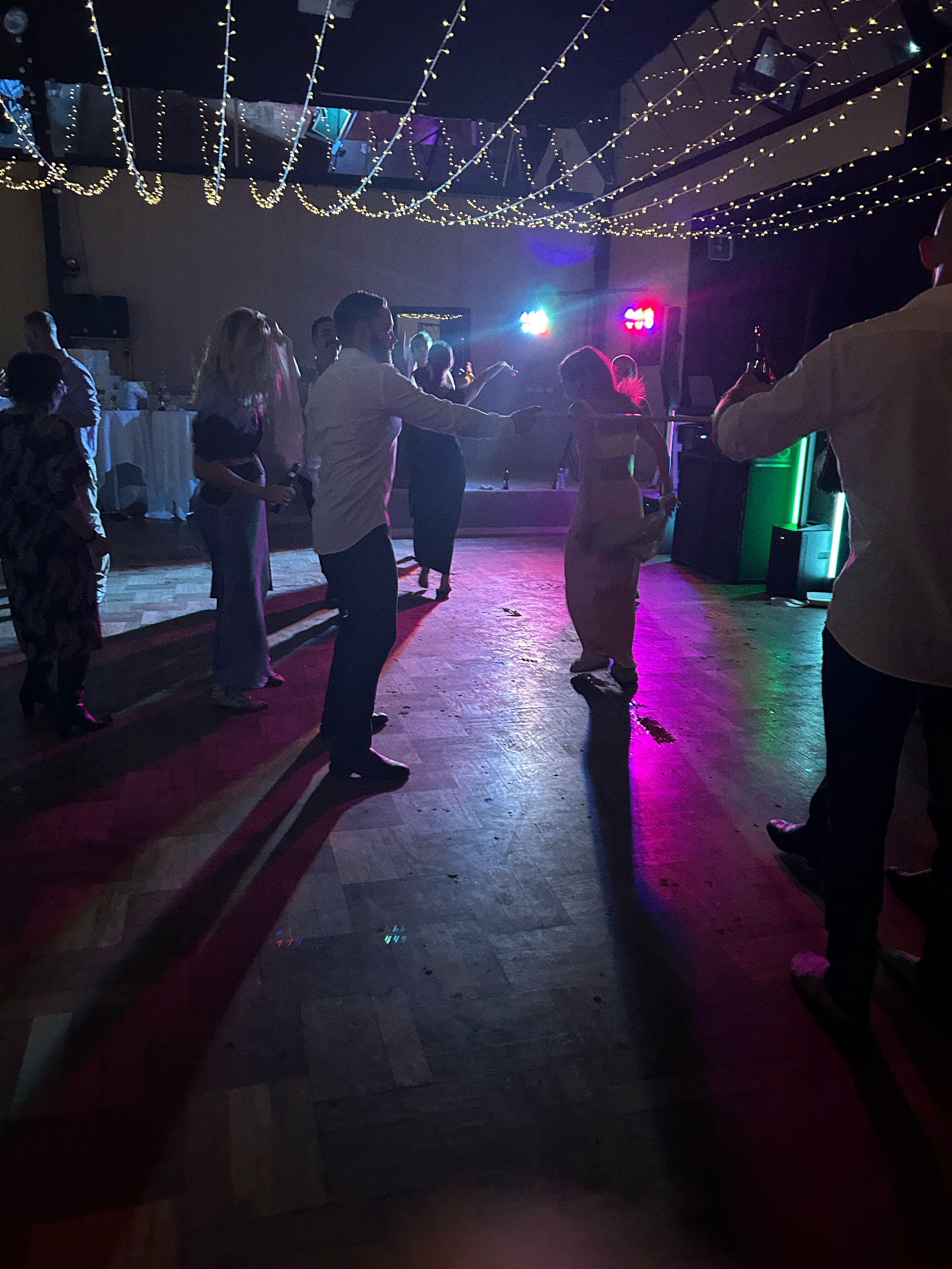 Et dansegulv med dansende mennesker, lyskæder hængt over hovederne og et sceneområde med et bord og stole.