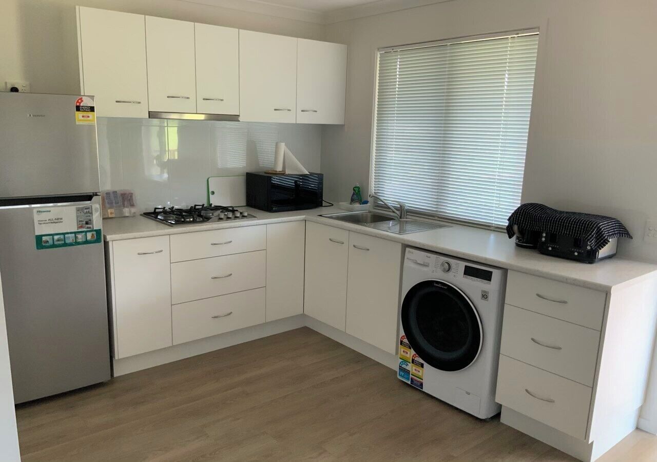 A modern kitchen with white cabinets, a stainless steel refrigerator, a gas stove, a washing machine, and a window with blinds.