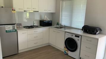 A modern kitchen with white cabinets, a stainless steel refrigerator, a gas stove, a washing machine, and a window with blinds.