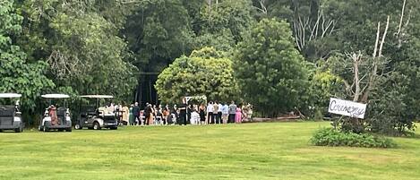 A group of people gathered outdoors, possibly at a ceremony, with golf carts and a sign indicating a ceremony.