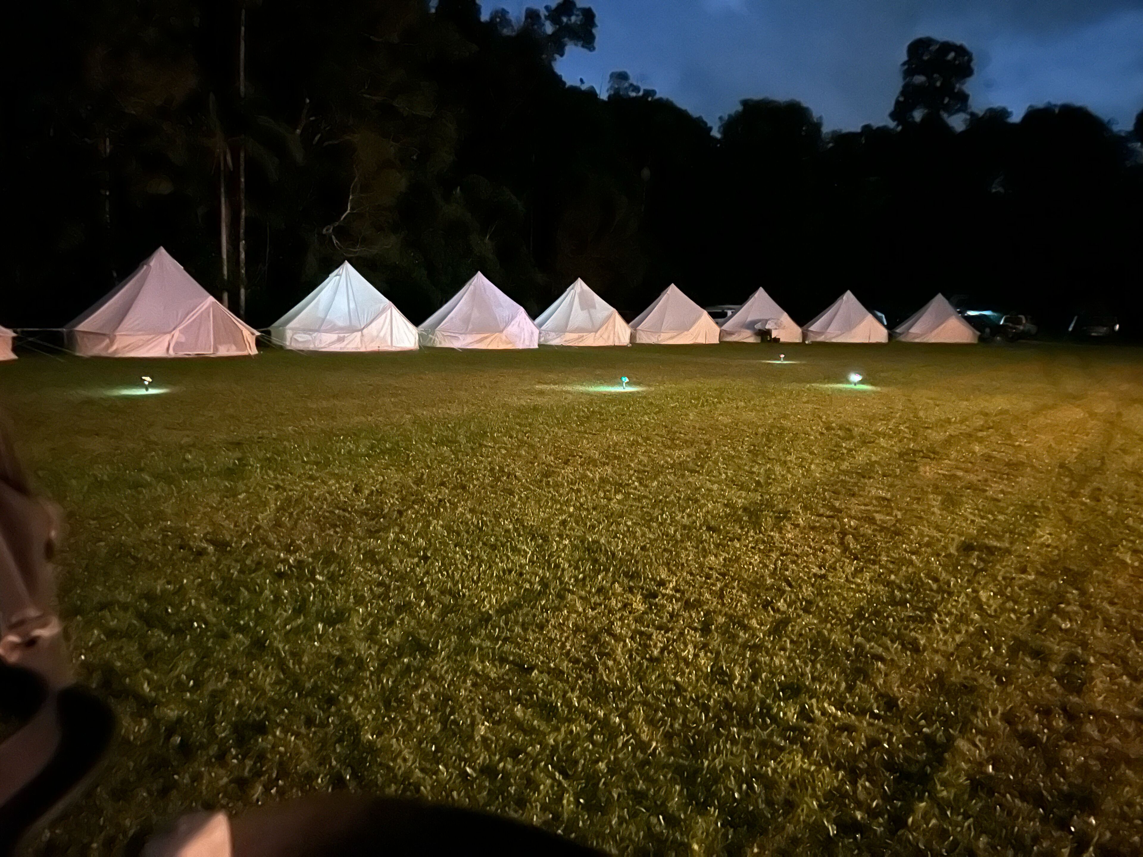 Une rangée de tentes blanches sur un champ d’herbe, la nuit, avec des lumières disposées au sol.