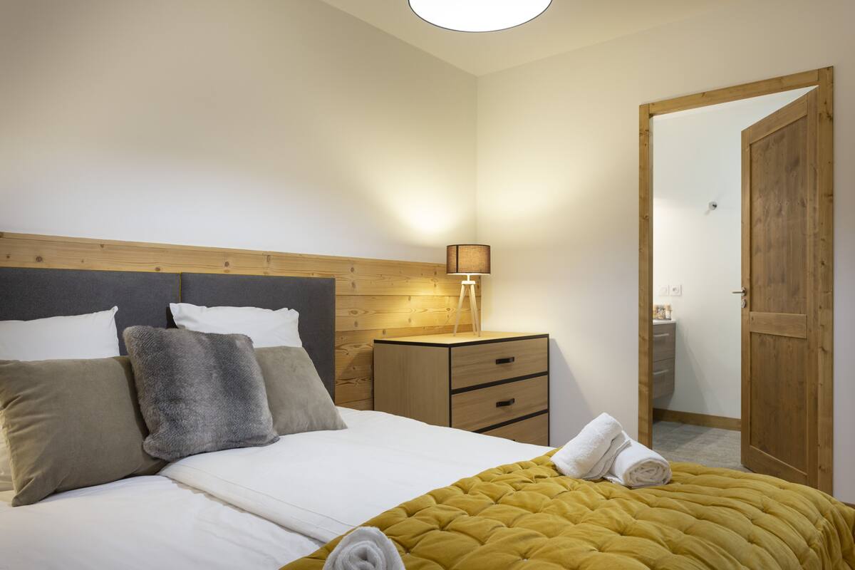 Ein ordentlich bezogenes Bett mit Kopfteil, ein Nachttisch mit Lampe und eine Holztür.