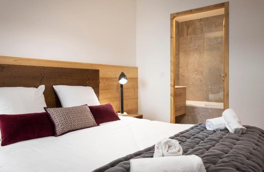 Habitación de hotel con cabecera de madera, cama con ropa de cama blanca y oscura, mesita de noche con lámpara y puerta de madera que da a un baño.