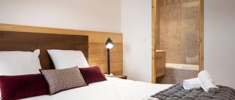 Habitación de hotel con cabecera de madera, cama con ropa de cama blanca y oscura, mesita de noche con lámpara y puerta de madera que da a un baño.