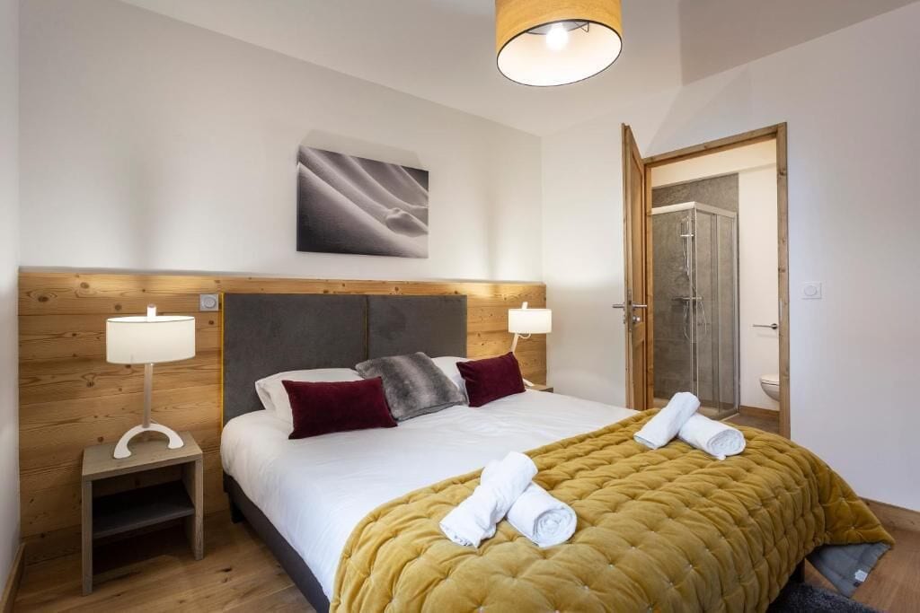 Habitación de hotel con cabecera de madera, cama con ropa de cama blanca, dos almohadas, colcha color mostaza, mesita de noche con lámpara y un cuadro en la pared.