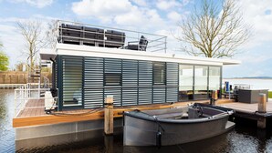 Une maison flottante moderne dotée d’une terrasse, d’un petit bateau amarré devant, et offrant une vue sur l’eau et le ciel.