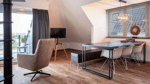 Una sala de estar moderna con un sofá, una mesa de comedor con sillas, un televisor sobre un trípode y un ventanal con persianas.