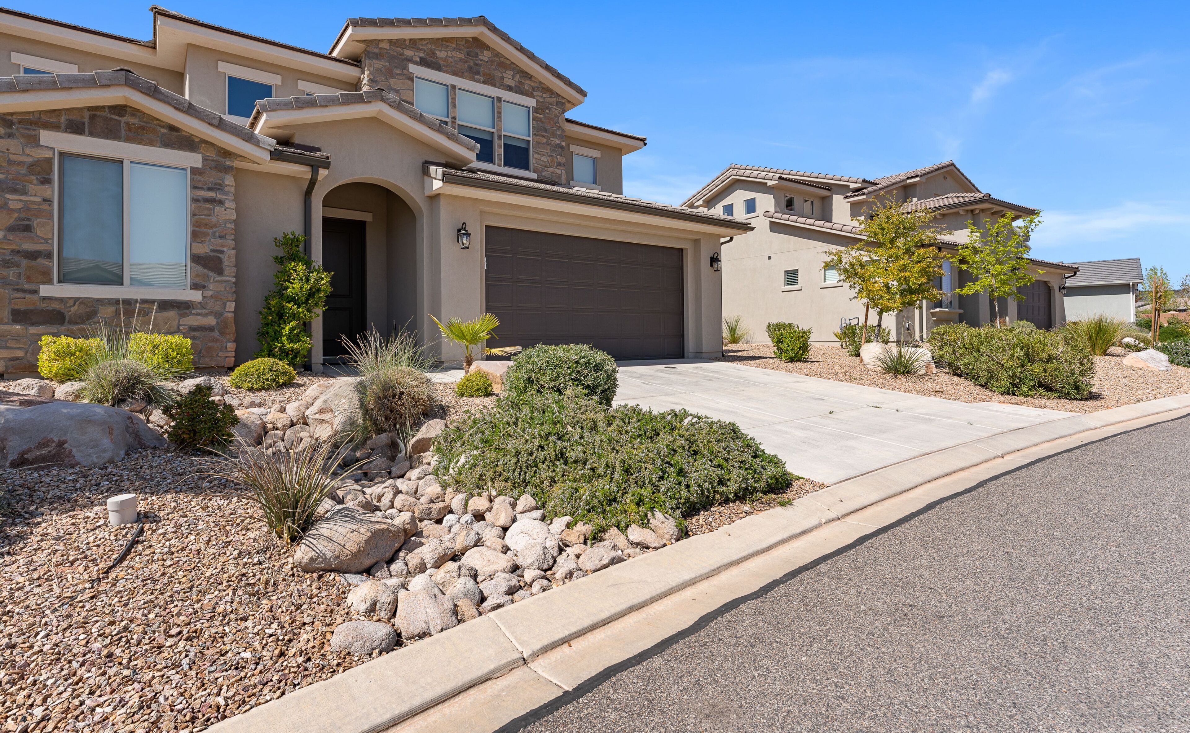 A modern two-story house with a stone facade, a garage, and a landscaped front yard with various shrubs and rocks.