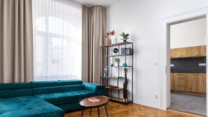 A modern living room with a teal sectional sofa, a wooden herringbone floor, a round coffee table, a black metal shelving unit with decorative items, and a view into a kitchen through an open door. - City Buzz Apartment - O2 Arena - Sleeps 6 (Prague)