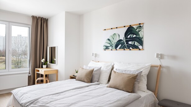 A modern bedroom with a large bed, a bedside table, a bench, and a window with curtains.