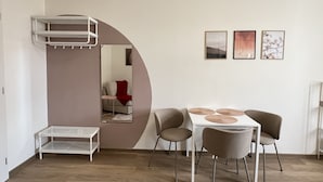 Un salon moderne avec un canapé blanc, une table à manger avec quatre chaises, une petite table d’appoint blanche et une étagère blanche. Trois tableaux encadrés sont accrochés au mur.